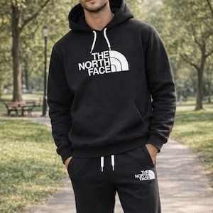 Puede incluir: Conjunto de sudadera con capucha y pantalón de chándal negros con el logo blanco "THE NORTH FACE". La sudadera tiene una capucha con cordón y un bolsillo delantero. El pantalón de chándal tiene cintura con cordón y el logo de la marca en la pierna izquierda.