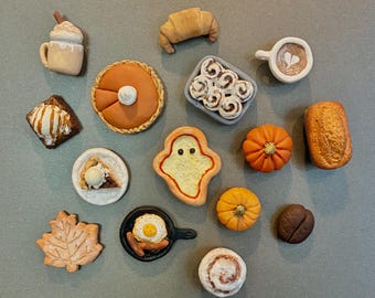 Puede incluir: Una colección de objetos en miniatura con temática de comida que incluye una taza con nata montada, un cruasán, una tarta de calabaza, una bandeja de rollitos de canela, una taza de café, un brownie con helado, una galleta con forma de fantasma, un pan, un trozo de tarta, dos calabazas, una galleta con forma de hoja, una sartén con un huevo y salchicha, un grano de café y un rollito de canela.