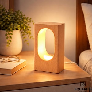 May include: A rectangular wooden table lamp with a glowing, oval-shaped cutout. The lamp is made of light-colored wood and sits on a wooden nightstand. A small plant in a white pot, a book, and glasses are also on the nightstand.