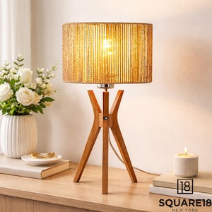 May include: A wooden tripod table lamp with a woven lampshade. The lampshade is a light brown color and the base is made of wood. The lamp is on a wooden table. The text "18 SQUARE18 NEW YORK" is visible.