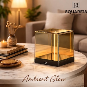 May include: A square, amber-colored lamp with a black base sits on a marble table. The lamp has a power button and the text "Ambient Glow" is at the bottom. The Square18 New York logo is in the top right corner.