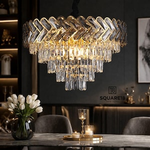 May include: A large, multi-tiered chandelier with a gold-colored frame and numerous crystal accents. The chandelier hangs above a dining table, with a vase of white flowers and candles visible. The text "SQUARE18 NEW YORK" is in the image.