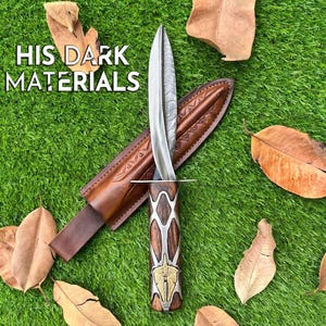Op de afbeelding: Een dolk van Damascus-staal met een houten handvat voorzien van zilverkleurige metalen overlays en een koperen inleg. De dolk bevat een bruine lederen schede met reliëfpatronen. De tekst "HIS DARK MATERIALS" is links zichtbaar.
