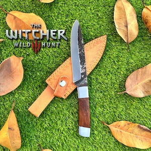 Puede incluir: Un cuchillo artesanal con mango de madera y hoja plateada, sobre una funda de cuero marrón claro. El cuchillo se encuentra sobre un fondo de hierba verde y hojas marrones dispersas. El texto "THE WITCHER WILD HUNT" es visible.