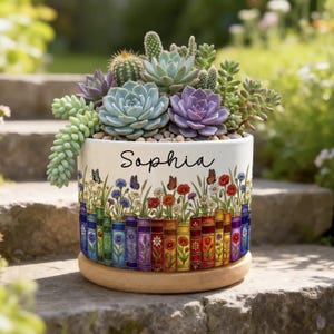 Könnte beinhalten: Ein weißer Keramik-Blumentopf mit einem farbenfrohen Blumen- und Buchdesign, gefüllt mit einer Vielzahl von Sukkulenten. Der Topf trägt den Namen "Sophia". Der Topf steht auf einem Holzsockel, ideal, um jedem Raum einen Hauch von Natur zu verleihen.