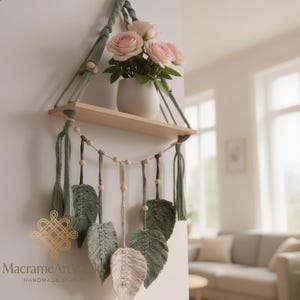 May include: A macrame wall shelf with a wooden shelf and decorative leaves. The shelf is suspended by green macrame rope and holds a white vase with pink roses. The shelf has a natural wood finish.
