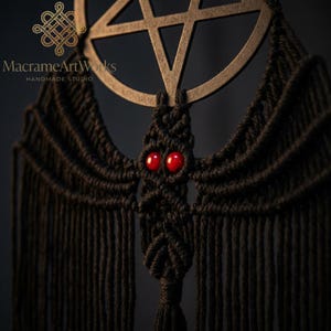 May include: Black macrame wall hanging featuring a wooden pentagram and a central bat-like figure with red bead eyes. The piece has a fringe at the bottom and is from MacrameArtWorks.
