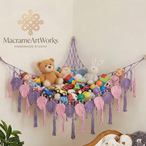 May include: A macrame toy hammock in a corner, filled with stuffed animals and toys. The hammock is purple with pink and purple leaf accents and tassels. The text "MacrameArtWorks HANDMADE STUDIO" is visible.