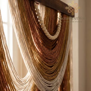 Puede incluir: Un tapiz de macramé hecho a mano con una barra de madera y hebras de hilo en crema, marrón y dorado. Una cadena de cuentas de madera añade un toque decorativo. El texto "Macrame ArtWork" es visible.