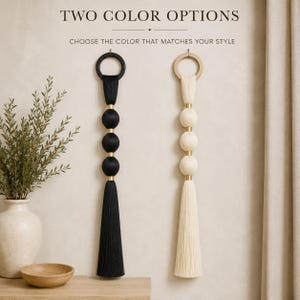 May include: Two wall hanging decorations featuring a circular top ring, three spherical ornaments, and a long tassel. One decoration is black, and the other is cream. Both feature gold-toned metal spacers between the spheres. Text reads TWO COLOR OPTIONS.