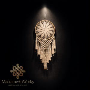 May include: A cream-colored macrame dream catcher with a floral design in the center. The dream catcher has a circular wooden frame and is adorned with hanging tassels. The logo "MacrameArtWorks" is visible at the bottom.