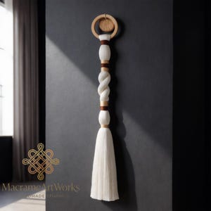 Puede incluir: Un colgante de pared de macramé hecho a mano con un anillo y detalles de madera. El diseño incorpora una cuerda blanca trenzada y una larga borla blanca. Los tonos marrones y de madera natural complementan las fibras blanquecinas. El logo "MacrameArtWorks" es visible.