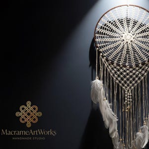 May include: A handcrafted dreamcatcher with a circular woven web design, featuring white feathers and wooden beads. The dreamcatcher is suspended against a dark blue background, with the text "MacrameArtWorks Handmade Studio" in the lower left corner.