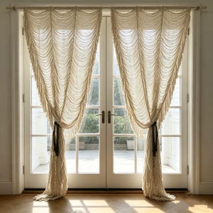 Puede incluir: Un par de cortinas de macramé color crema colgadas de una barra horizontal sobre una puerta doble de cristal. Las cortinas presentan intrincados patrones de cordones anudados que caen en festones, recogidas abajo con lazos oscuros.