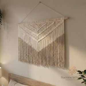 Puede incluir: Un tapiz de pared de macramé beige con un diseño geométrico. La obra de arte está suspendida de un pasador de madera y una cuerda blanca. La parte inferior del macramé tiene flecos largos y fluidos. El texto "MacrameArtWorks Handmade Studio" es visible.