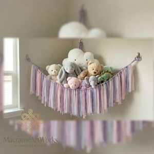 Puede incluir: Un macramé de pared en colores pastel, que sostiene varios peluches. El estante tipo hamaca presenta flecos rosas, morados y azules. Una almohada blanca en forma de nube está encima del estante. El texto "MacrameArtWorks" está en la parte inferior.