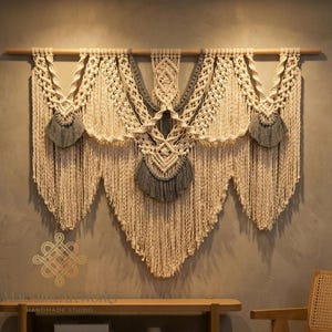 May include: A large macrame wall hanging in natural and gray tones. The handmade textile features intricate knotting patterns, including cascading fringe and decorative accents. The piece is suspended from a wooden dowel, creating a bohemian-inspired decor element.