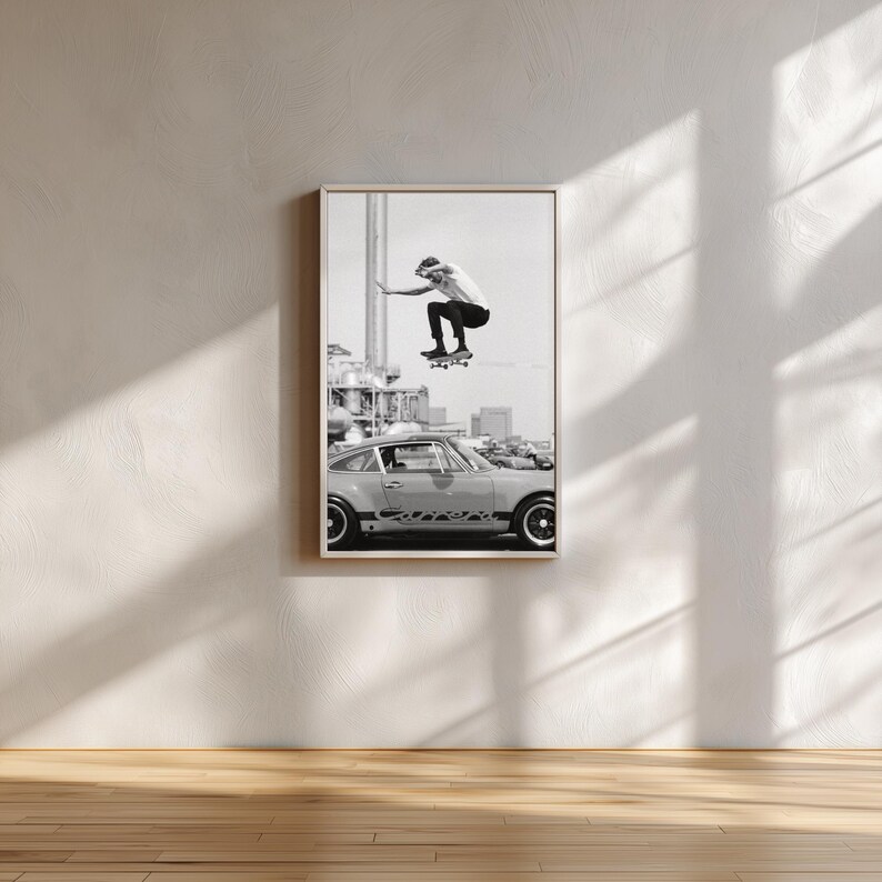 May include: Black and white framed print featuring a skateboarder mid-air above a classic car. The image captures a moment of action and urban culture, with a cityscape backdrop. The print is displayed on a white wall with natural light.