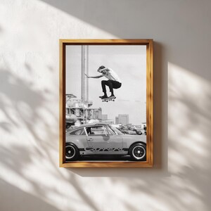 May include: Black and white framed print featuring a skateboarder mid-air above a vintage car. The print has a retro aesthetic, with the skateboarder wearing a white t-shirt and black pants. The car is a light color with black wheels.