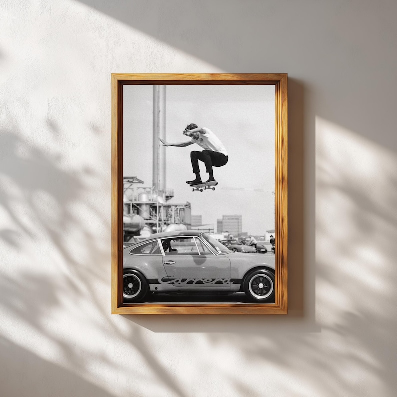 May include: Black and white framed print featuring a skateboarder mid-air above a vintage car. The print has a retro aesthetic, with the skateboarder wearing a white t-shirt and black pants. The car is a light color with black wheels.