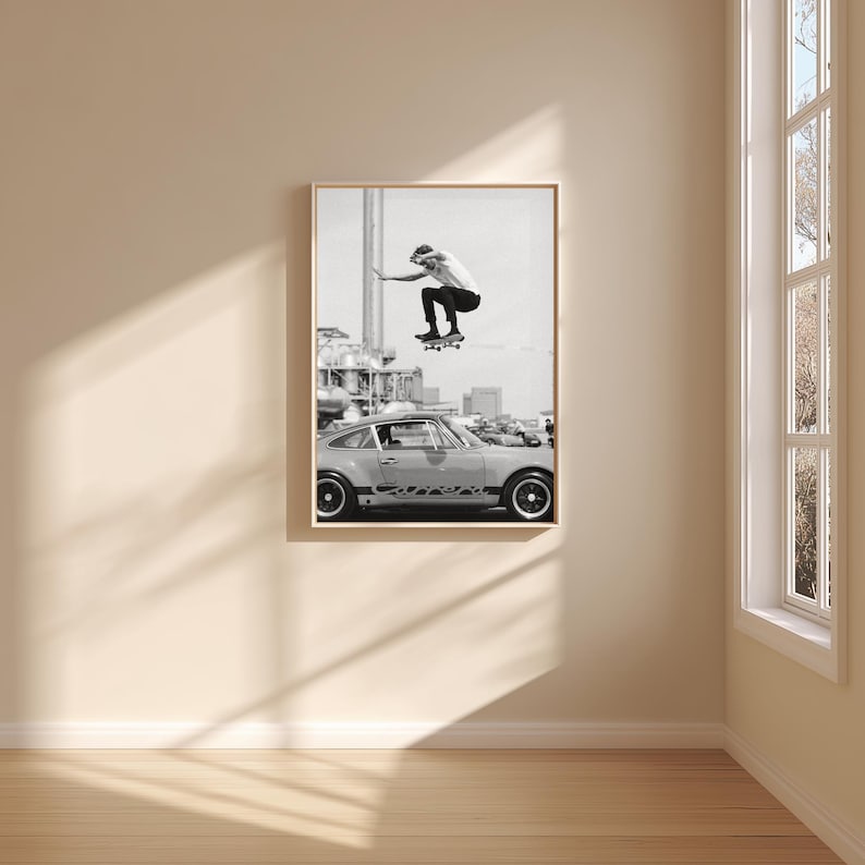 May include: Black and white framed print featuring a skateboarder mid-air above a vintage sports car. The image captures a moment of action and urban culture, with a focus on the skateboarder's athleticism and the car's classic design. The print is displayed in a room with natural light.
