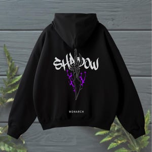 Op de afbeelding: Zwarte hoodie met het woord "SHADOW" in witte, gestileerde letters en een paarse en witte zwaardafbeelding. Het woord "MONARCH" is onder de afbeelding gedrukt.