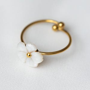 May include: A gold-toned ring featuring a thin, open-ended band with two small spheres at the ends. The ring is adorned with a white, five-petaled flower ornament that has a small gold-toned center, creating a delicate and simple jewelry piece.