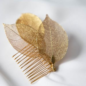 May include: A gold-toned hair comb featuring three delicate, skeletonized leaf ornaments attached to the top. The leaves display a fine, intricate vein structure and are secured to the metallic comb base with thin, wrapped gold-colored wire.