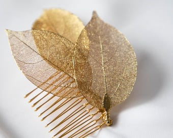 May include: A gold-toned hair comb featuring three delicate, skeletonized leaf ornaments attached to the top. The leaves display a fine, intricate vein structure and are secured to the metallic comb base with thin, wrapped gold-colored wire.