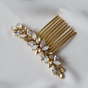 May include: A gold-toned hair comb with a decorative leaf design. The leaves are adorned with clear and opalescent crystals. The comb is set against a white, textured background, suggesting a bridal or formal accessory.