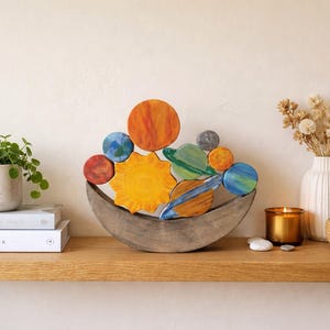 May include: A wooden solar system sculpture with planets and a sun in a crescent-shaped base. The planets are painted in various colors, including blue, orange, and green. A small potted plant, books, a candle, and a vase with dried flowers are also on the shelf.