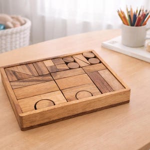 May include: A wooden puzzle with various geometric shapes, including rectangles, circles, and triangles, arranged within a square wooden frame. The puzzle is made of light brown wood and sits on a light-colored wooden table. A white mug with colored pencils is in the background.