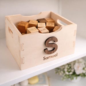 May include: A wooden toy box filled with wooden alphabet blocks. The box is light-colored with a dark wooden letter "S" and the name "Samuel" on the front. The blocks are engraved with letters and symbols.