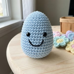 Puede incluir: Un juguete de peluche en forma de huevo, de color azul claro, tejido a ganchillo, con una cara sonriente. El juguete tiene ojos negros de botón y una boca negra cosida. Cerca hay flores de ganchillo de colores pastel, lo que sugiere un tema primaveral o de Pascua.