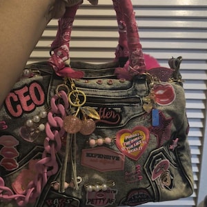 Op de afbeelding: Een denim handtas met roze accenten en versieringen. De tas heeft het woord "CEO" en andere patches. Het heeft een ritssluiting, roze ketting en decoratieve bedels. De handvatten zijn omwikkeld met roze stof.