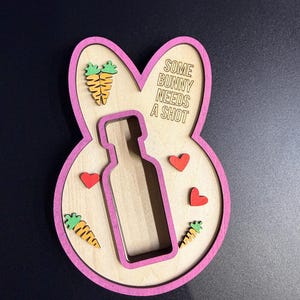 May include: A wooden bunny-shaped plaque with a pink border, featuring a central cutout for a bottle. The plaque includes the text 'SOME BUNNY NEEDS A SHOT' and is decorated with three small red wooden hearts and three orange carrots with green tops.
