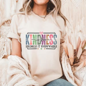 May include: A beige t-shirt with the words "KINDNESS is free, SPRINKLE IT EVERYWHERE" in colorful letters. The design includes a pink heart and star accents. The shirt is made of a soft material.