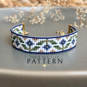 May include: A beaded loom bracelet featuring a floral design in white, blue, green, and gold. It has a dark blue border and gold clasps. The text "LOOM BRACELET PATTERN" is displayed below the bracelet.