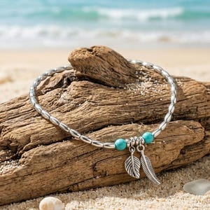 May include: Silver beaded anklet with turquoise accents and feather charms. The anklet is displayed on a piece of driftwood on a sandy beach, with the ocean in the background. A delicate piece of jewelry for a beachy look.