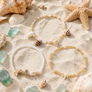 May include: Three beaded bracelets featuring small white and gold-toned beads. One bracelet includes brown and white seashell charms, another features a cluster of small seashells, and the third is accented with a single white starfish charm.