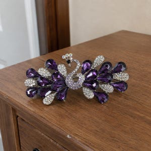 May include: A silver-toned hair clip shaped like a peacock, featuring deep purple teardrop-shaped crystals and smaller clear rhinestones. The peacock design is centered with a curved neck and a fanned tail made of alternating purple and clear stones.