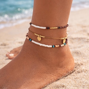 May include: Three beaded anklets in a beach setting. The top anklet features white and brown beads with gold accents. The middle anklet is gold with a small gold charm. The bottom anklet has white, brown, and turquoise beads.