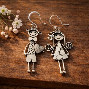 May include: A pair of silver-toned dangle earrings featuring stylized figures. One earring depicts a figure with a bow tie and a textured heart, while the other shows a figure with long hair and a polka-dot dress. Both have hook-style backings.
