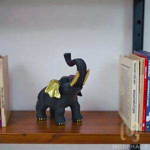 Puede incluir: Una figura de elefante negro con detalles dorados en las orejas, colmillos y patas. La trompa está levantada y la figura se exhibe en una estantería de madera. Se ven libros a ambos lados del elefante.