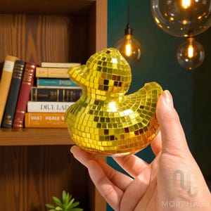 May include: A golden mosaic duck figurine. The duck is covered in small, reflective, square tiles, creating a shimmering effect. The figurine is held in a hand, with a bookshelf and decorative lights in the background.
