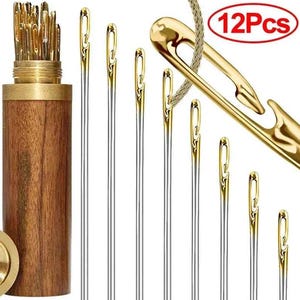 May include: A set of 12 sewing needles with gold-toned eyes and silver shafts. The needles are displayed with a wooden storage container and a large gold-toned needle. The text "12Pcs" is in a red circle.