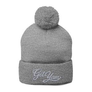 May include: A grey beanie hat with a matching pom-pom. The folded brim displays the text "Get Yum" in a cursive, rhinestone-style font. The hat is made of a soft, knitted material.