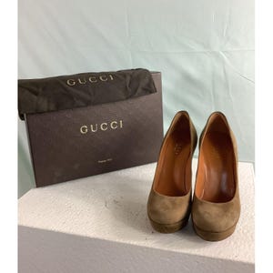 May include: A pair of olive green suede platform pumps with rounded toes and high heels, accompanied by a dark brown Gucci branded box and a matching fabric dust bag with the Gucci logo printed in gold lettering on the front.