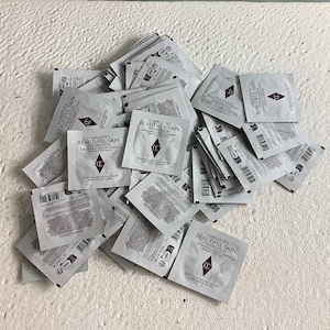 May include: A large collection of square, white Charlotte's Beautiful Skin Foundation sample packets. Each individual 1 ml sachet features the brand logo, product name, and text in black, with various shade labels printed on the back of the packets.