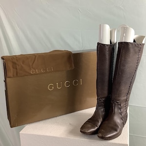 May include: A pair of dark brown knee-high boots with visible whipstitch detailing along the sides and white plastic boot trees inside. The boots are positioned next to a rectangular brown Gucci branded box and a matching brown fabric dust bag.